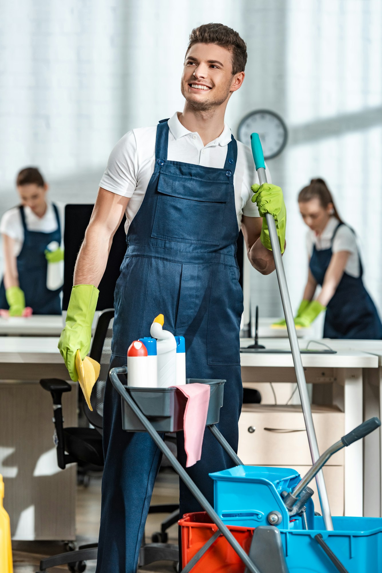 smiling cleaner standing near cart with cleaning supplies and looking away